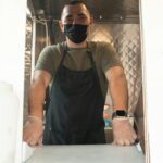 A masked food truck vendor ready to take orders at the window, offering a safe and welcoming service.